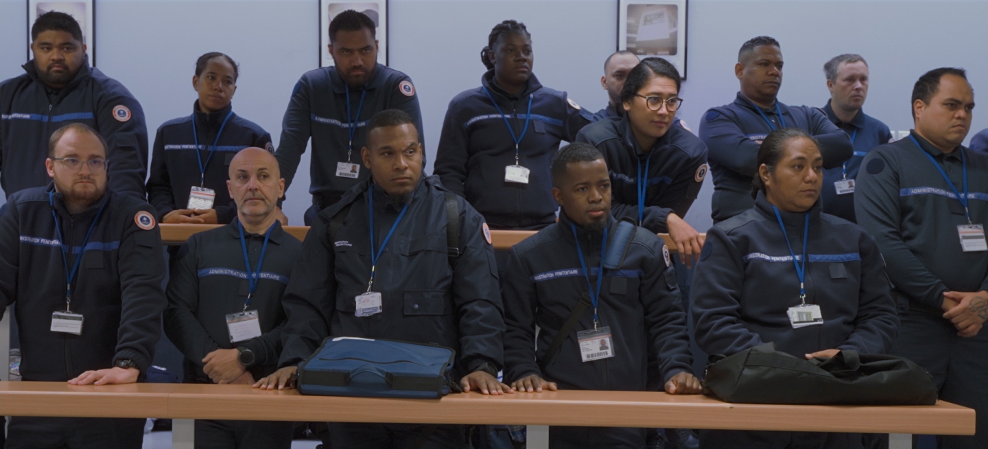 A group of adult students in a prison guard classroom stand attentively while listening to a speaker outside the frame. The racially diverse group of students are all wearing the same navy blue uniform with a white badge on a lanyard around their necks.