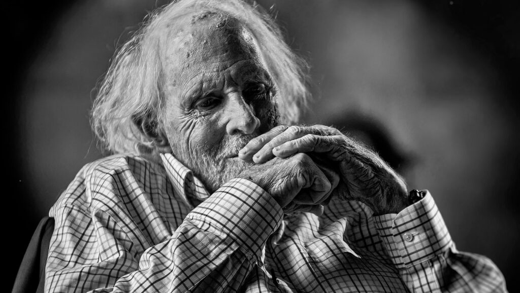 A black and white photo of actor Bruce Dern. He is a white man in his 80s with shaggy white hair. He is smiling and his seated with his hands raised to his chin.