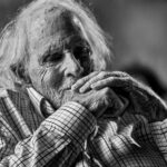 A black and white photo of actor Bruce Dern. He is a white man in his 80s with shaggy white hair. He is smiling and his seated with his hands raised to his chin.