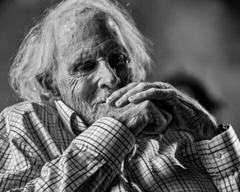 A black and white photo of actor Bruce Dern. He is a white man in his 80s with shaggy white hair. He is smiling and his seated with his hands raised to his chin.