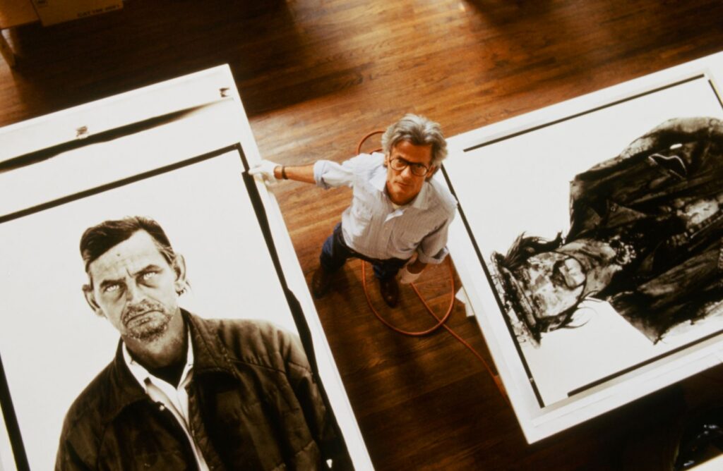 A man stands on a hardwood floor looking up towards the camera, which captures a view from above. There are two oversized black and white photographers on either side of him depicting portraits of working class men.