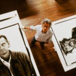 A man stands on a hardwood floor looking up towards the camera, which captures a view from above. There are two oversized black and white photographers on either side of him depicting portraits of working class men.