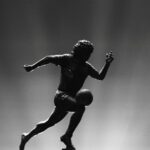 A silhouette of a male soccer player running after a ball appears in black and white. There are flood lights coming up from behind him.