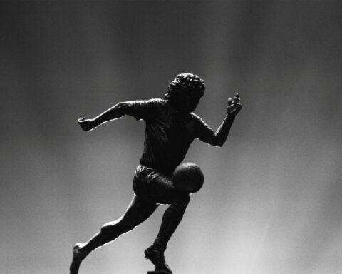 A silhouette of a male soccer player running after a ball appears in black and white. There are flood lights coming up from behind him.