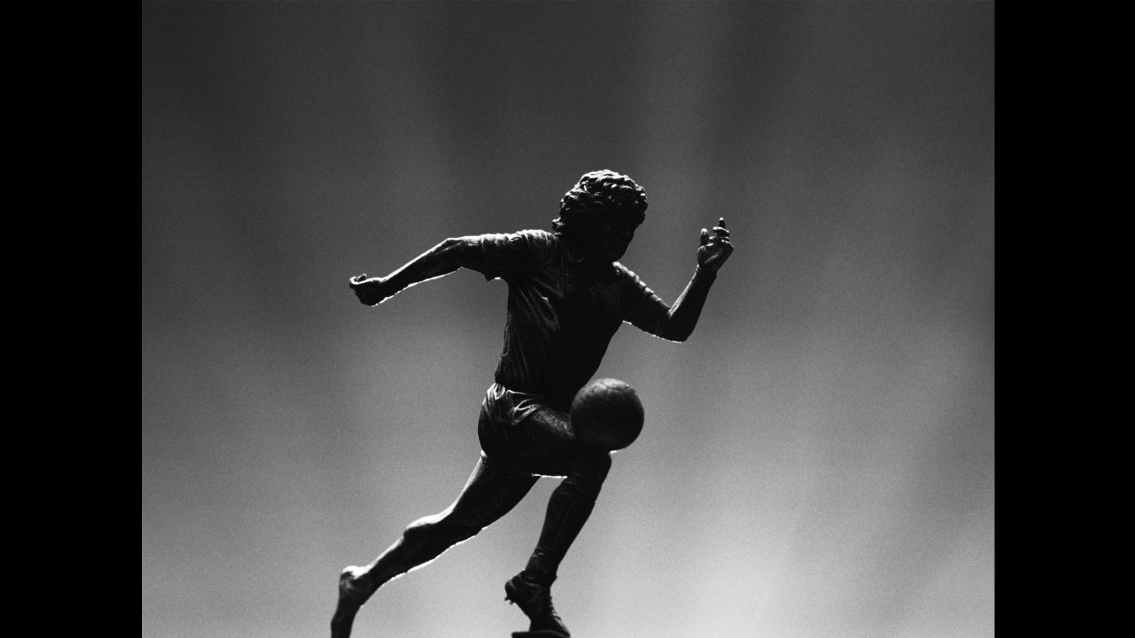 A silhouette of a male soccer player running after a ball appears in black and white. There are flood lights coming up from behind him.