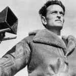 A black and white photo depicts British filmmaker David Lean standing in front of a camera. He is shot from a low angle, looking beyond the frame. He is a white man with brown hair, and he is wearing an overcoat.