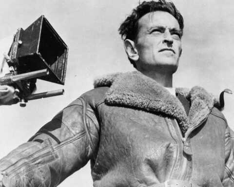 A black and white photo depicts British filmmaker David Lean standing in front of a camera. He is shot from a low angle, looking beyond the frame. He is a white man with brown hair, and he is wearing an overcoat.