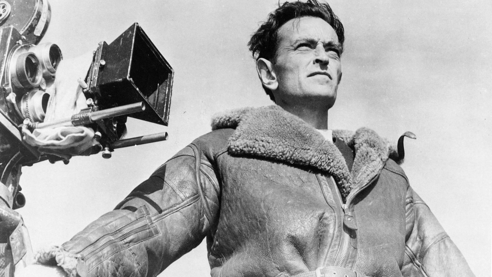 A black and white photo depicts British filmmaker David Lean standing in front of a camera. He is shot from a low angle, looking beyond the frame. He is a white man with brown hair, and he is wearing an overcoat.