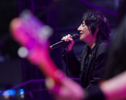 Rocker Carole Pope is views in profile as she sings on stage. A guitarist is in the foreground of the picture.