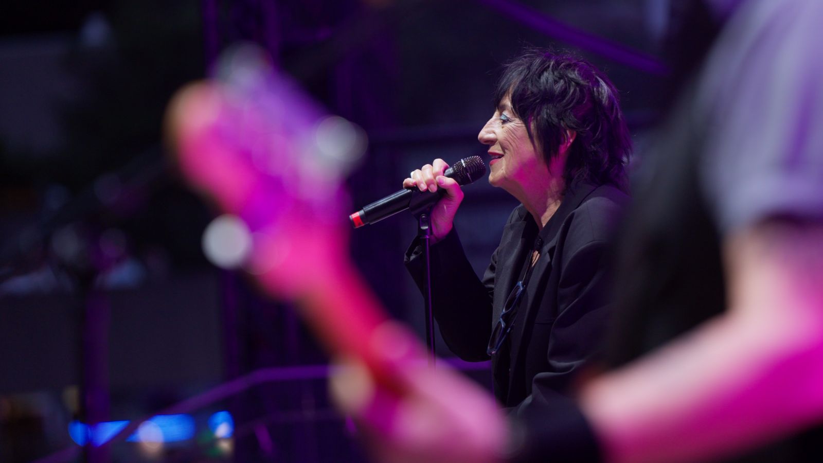 Rocker Carole Pope is views in profile as she sings on stage. A guitarist is in the foreground of the picture.