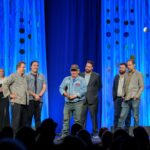 Six men accept an award on a stage with dark blue curtains behind them and two large trophy statues framing the stage.