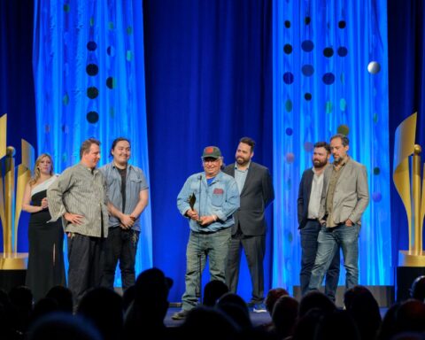 Six men accept an award on a stage with dark blue curtains behind them and two large trophy statues framing the stage.