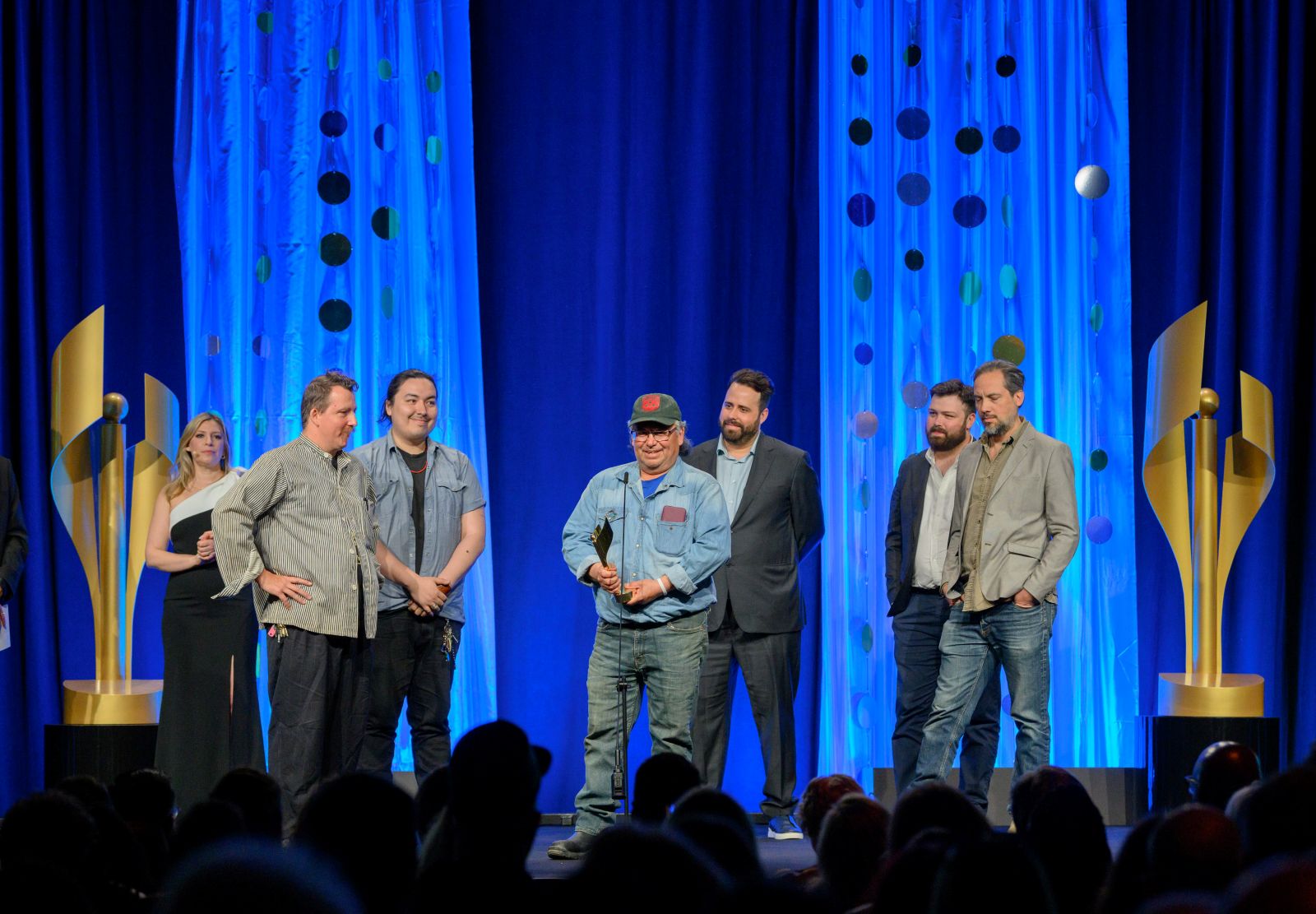 Six men accept an award on a stage with dark blue curtains behind them and two large trophy statues framing the stage.