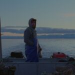 An oyster farmer stands in a boat at dusk.