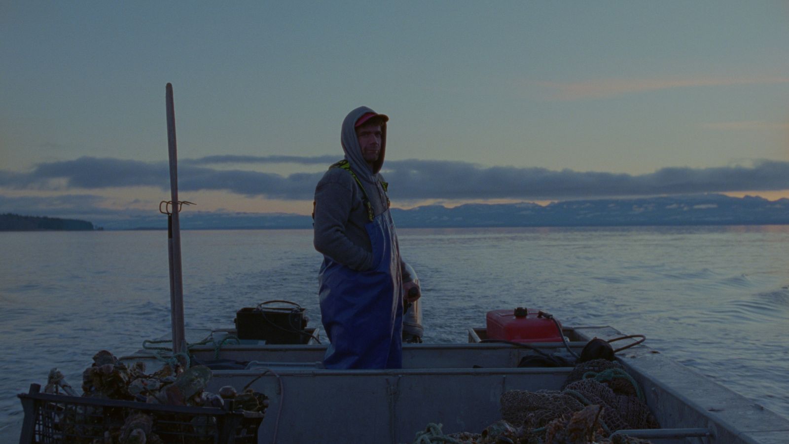 An oyster farmer stands in a boat at dusk.