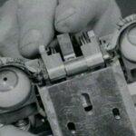 An archival image of a pair of hands touching an early robot. The machine has a pair of eyes.