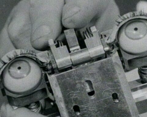 An archival image of a pair of hands touching an early robot. The machine has a pair of eyes.