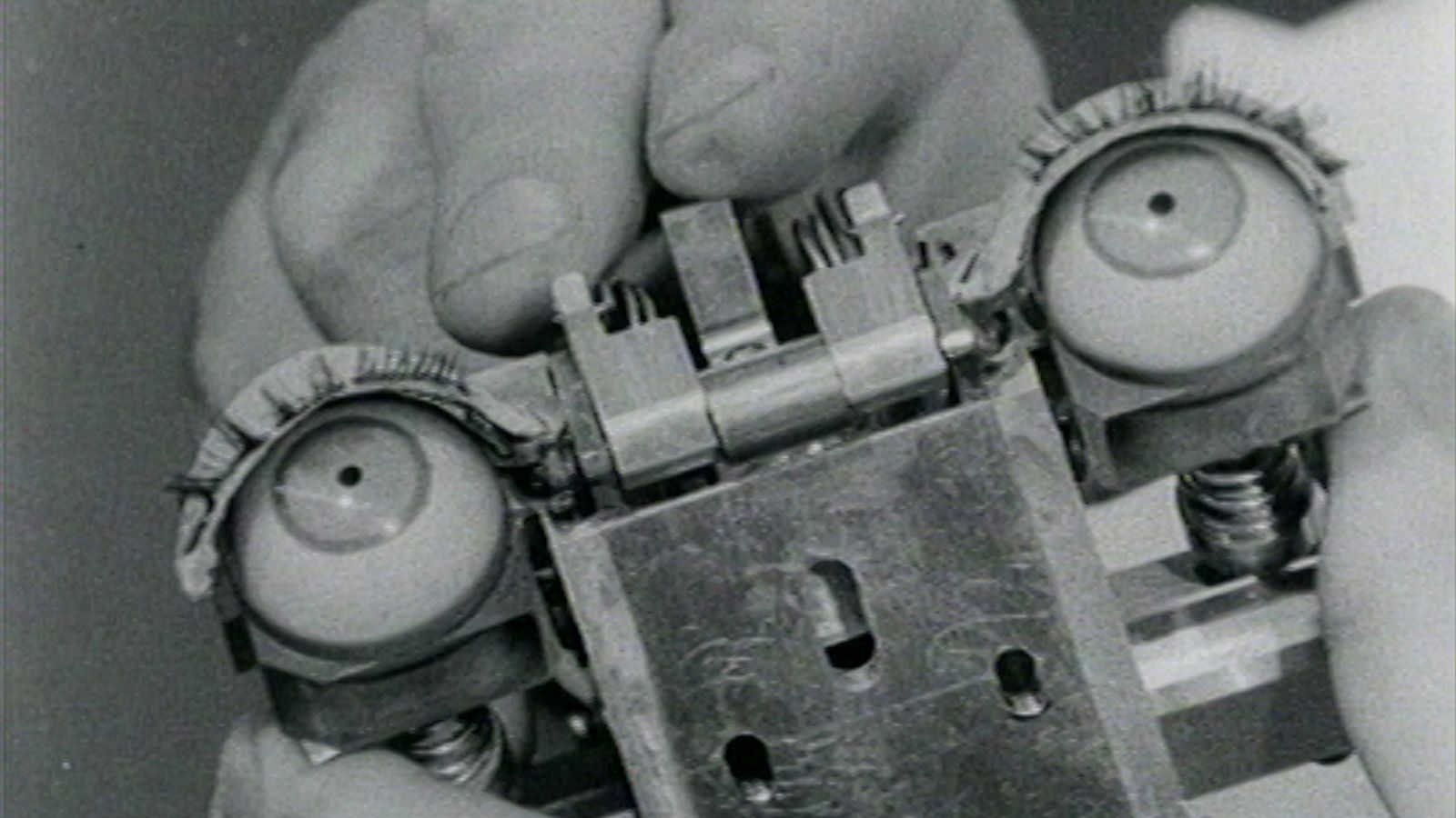 An archival image of a pair of hands touching an early robot. The machine has a pair of eyes.