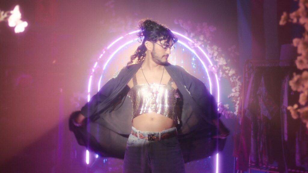 Bred D'Souza models a silver halter top wth a black cape. They have long dark hair and a bear and moustache, and stand in front of an archway made of pink neon lights.