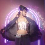 Bred D'Souza models a silver halter top wth a black cape. They have long dark hair and a bear and moustache, and stand in front of an archway made of pink neon lights.