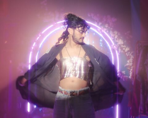 Bred D'Souza models a silver halter top wth a black cape. They have long dark hair and a bear and moustache, and stand in front of an archway made of pink neon lights.