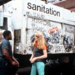 A blonde woman in blue jeans and an orange shirt with a black vest speaks with a male sanitation worker. He is Black and wearing a blue uniform. They are standing in front of a white garbage truck with Sanitation in black letters near the top of the vehicle. The truck is covered in graffiti.