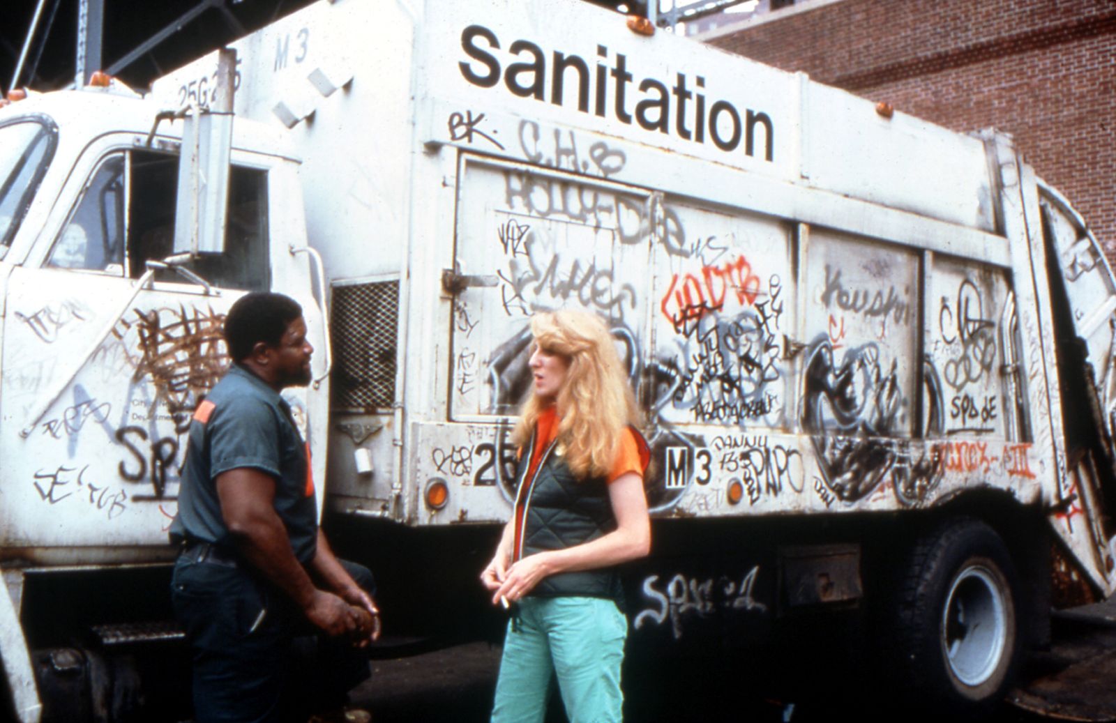 A blonde woman in blue jeans and an orange shirt with a black vest speaks with a male sanitation worker. He is Black and wearing a blue uniform. They are standing in front of a white garbage truck with Sanitation in black letters near the top of the vehicle. The truck is covered in graffiti.