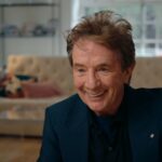 Actor Martin Short is a white man in his 70s with red hair. He is seated for an interview, smiling and leaning in towards the camera, and wearing a dark suit.
