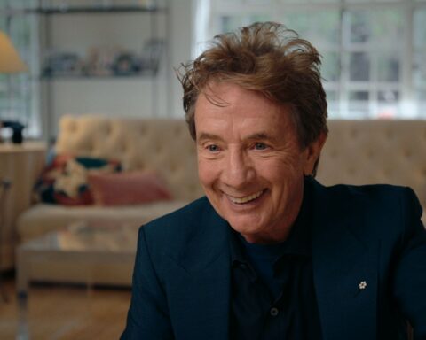 Actor Martin Short is a white man in his 70s with red hair. He is seated for an interview, smiling and leaning in towards the camera, and wearing a dark suit.