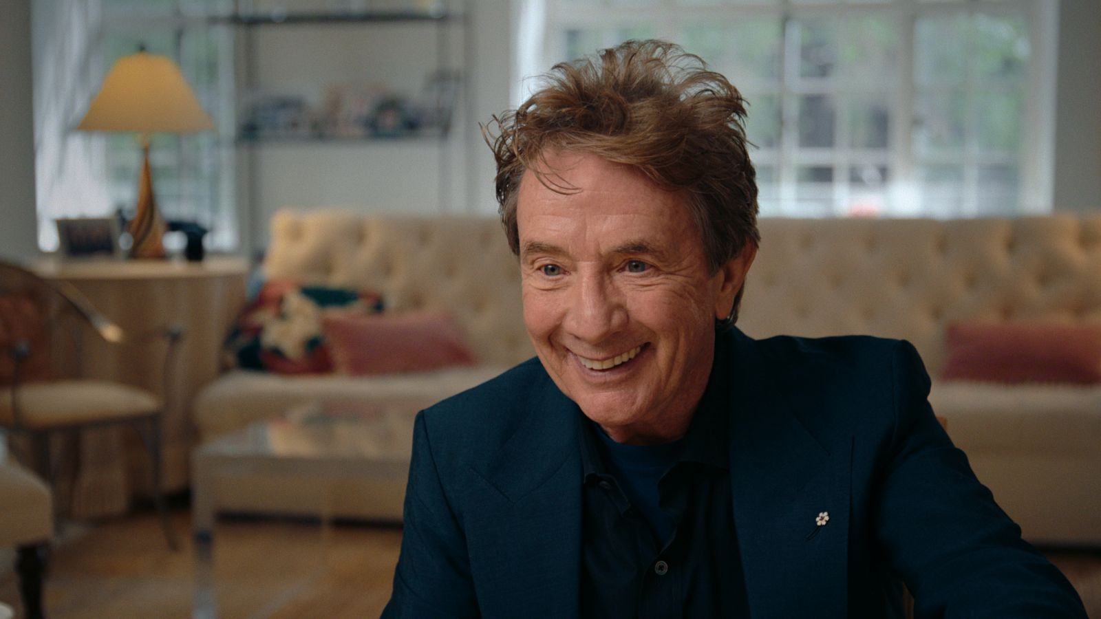 Actor Martin Short is a white man in his 70s with red hair. He is seated for an interview, smiling and leaning in towards the camera, and wearing a dark suit.
