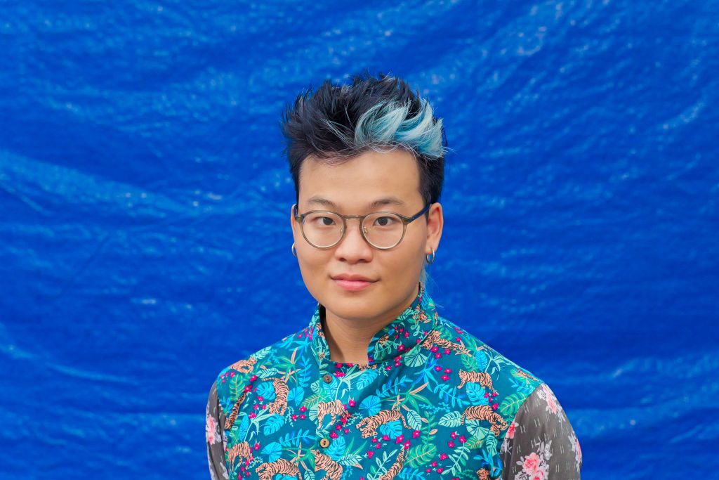 Nhật Quang Nông is a Vietnamese filmmaker in his late 20s. He has glasses and a blue streak in his black hair. He is wearing a blue, pink, and purple patterned robe, and is pictured in front of a blue backdrop.