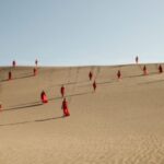 Women in red dresses walk down a sand dune.