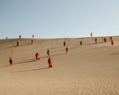 Women in red dresses walk down a sand dune.