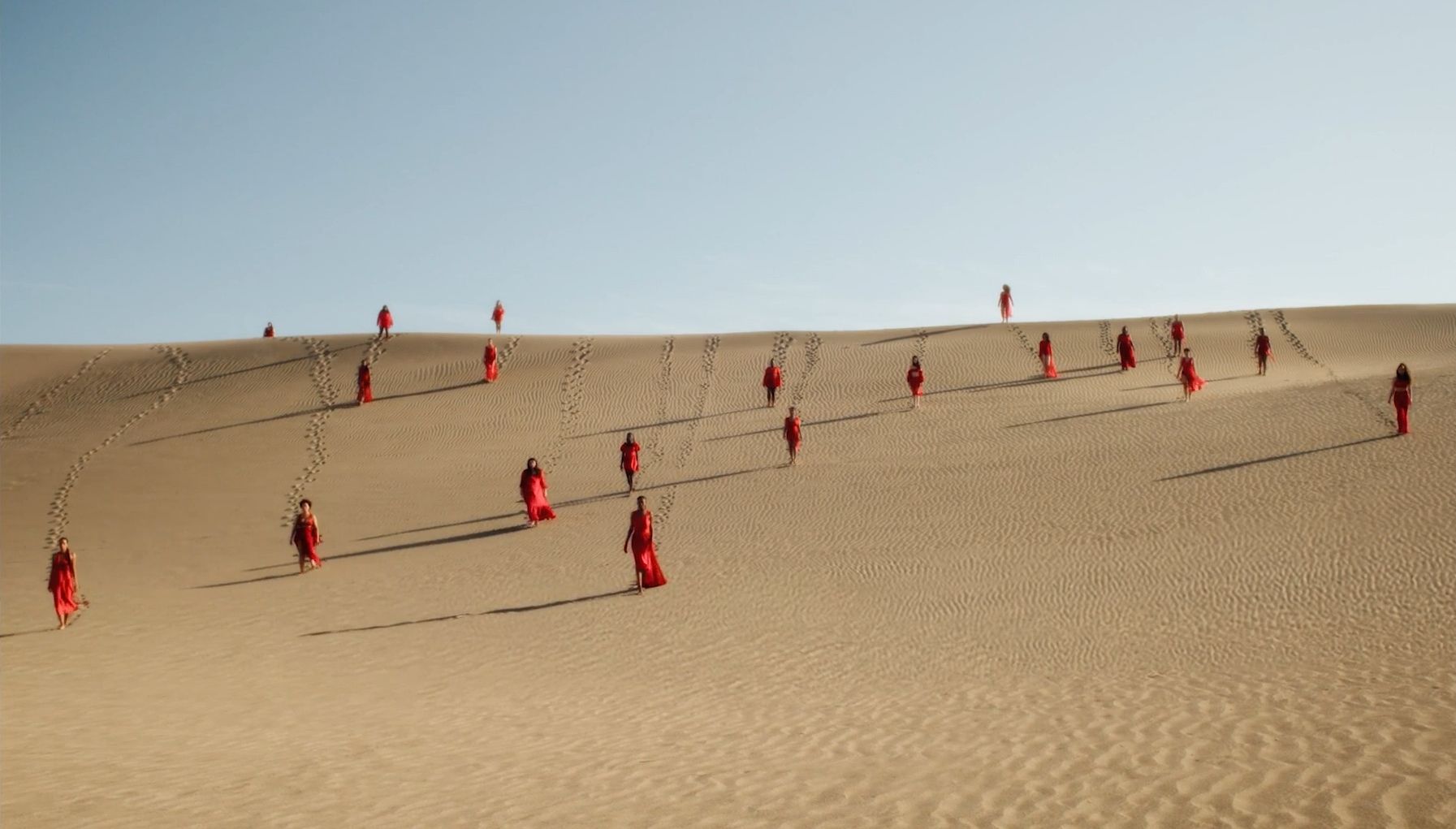 Women in red dresses walk down a sand dune.