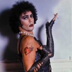 Actor Tim Curry appears as Dr. Frank-N-Furter in The Rocky Horror Picture Show, as seen in the documentary Strange Journey. He is a young white man in a brown curly-haired wig, posing with his hand raised holding a cigarette, while wearing a black corset, black arm-length globes, a pearl necklace, and heavy make-up.