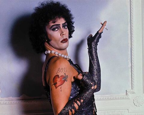 Actor Tim Curry appears as Dr. Frank-N-Furter in The Rocky Horror Picture Show, as seen in the documentary Strange Journey. He is a young white man in a brown curly-haired wig, posing with his hand raised holding a cigarette, while wearing a black corset, black arm-length globes, a pearl necklace, and heavy make-up.