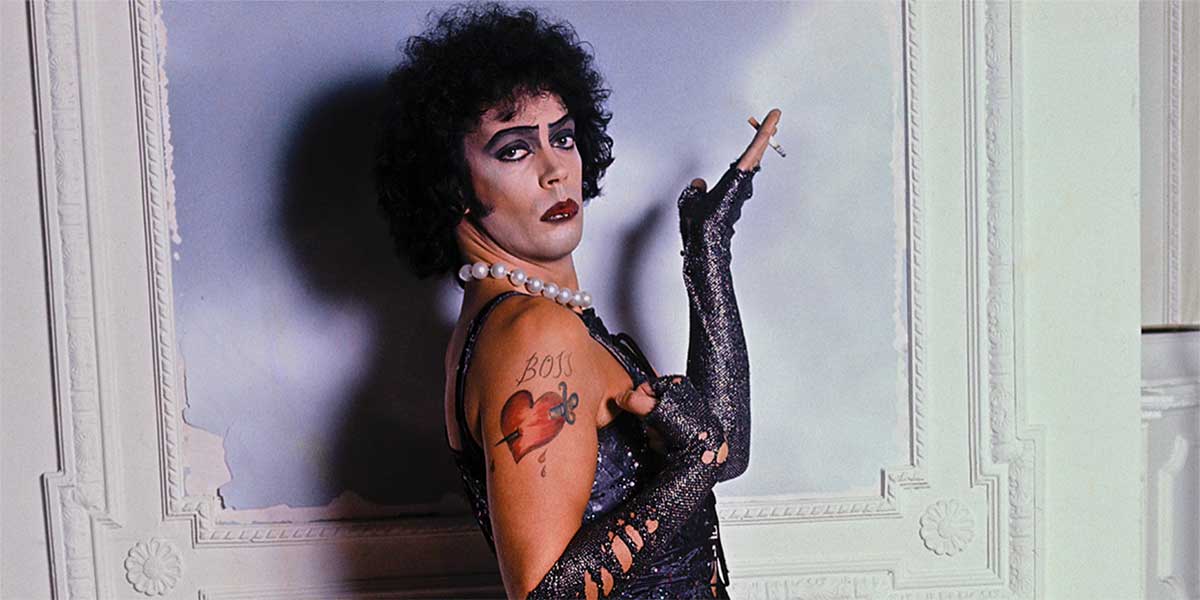 Actor Tim Curry appears as Dr. Frank-N-Furter in The Rocky Horror Picture Show, as seen in the documentary Strange Journey. He is a young white man in a brown curly-haired wig, posing with his hand raised holding a cigarette, while wearing a black corset, black arm-length globes, a pearl necklace, and heavy make-up.