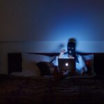 A woman in a black ski mask sits on a bod, reading a laptop. Her room is dark and she is illuminated by the light of the computer screen.