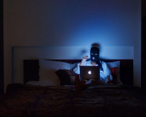 A woman in a black ski mask sits on a bod, reading a laptop. Her room is dark and she is illuminated by the light of the computer screen.