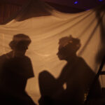Two filmmakers are pictured in silhouette. They are sitting in a blanket fort and having a conversation.