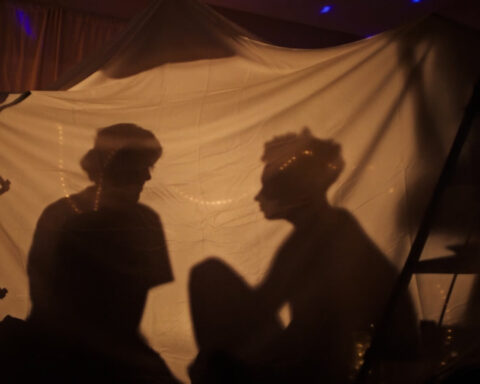 Two filmmakers are pictured in silhouette. They are sitting in a blanket fort and having a conversation.