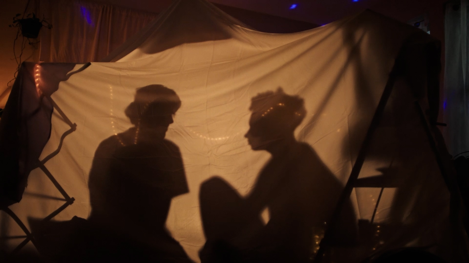 Two filmmakers are pictured in silhouette. They are sitting in a blanket fort and having a conversation.