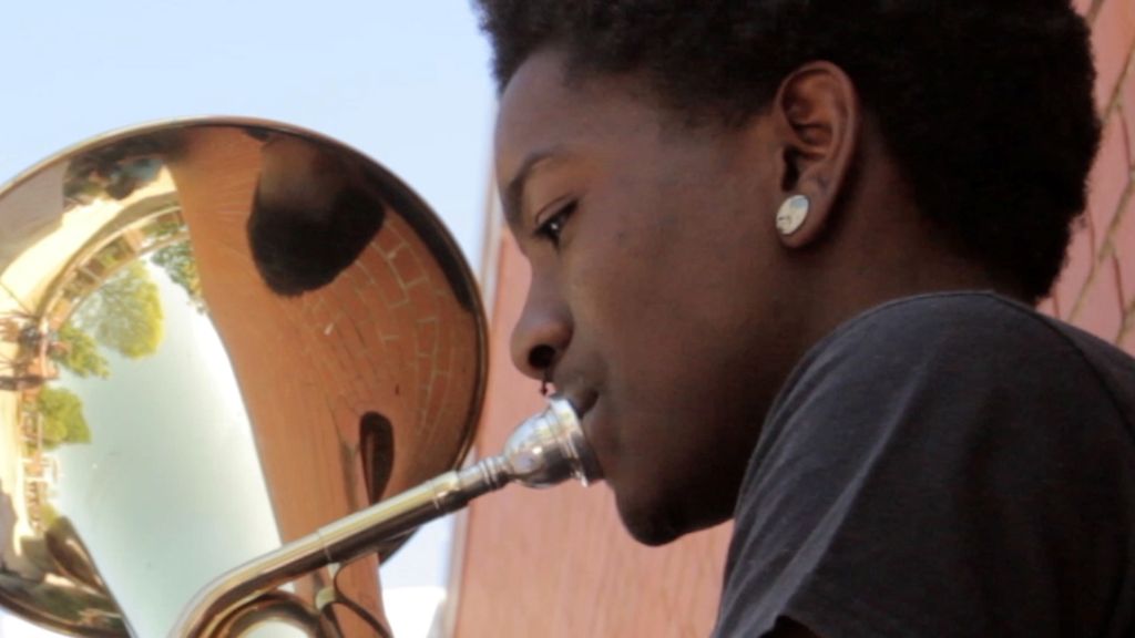 Blake, a Black teenager, plays a tuba. He is pictured in close-up and is wearing a black t-shirt and diamond wearing.
