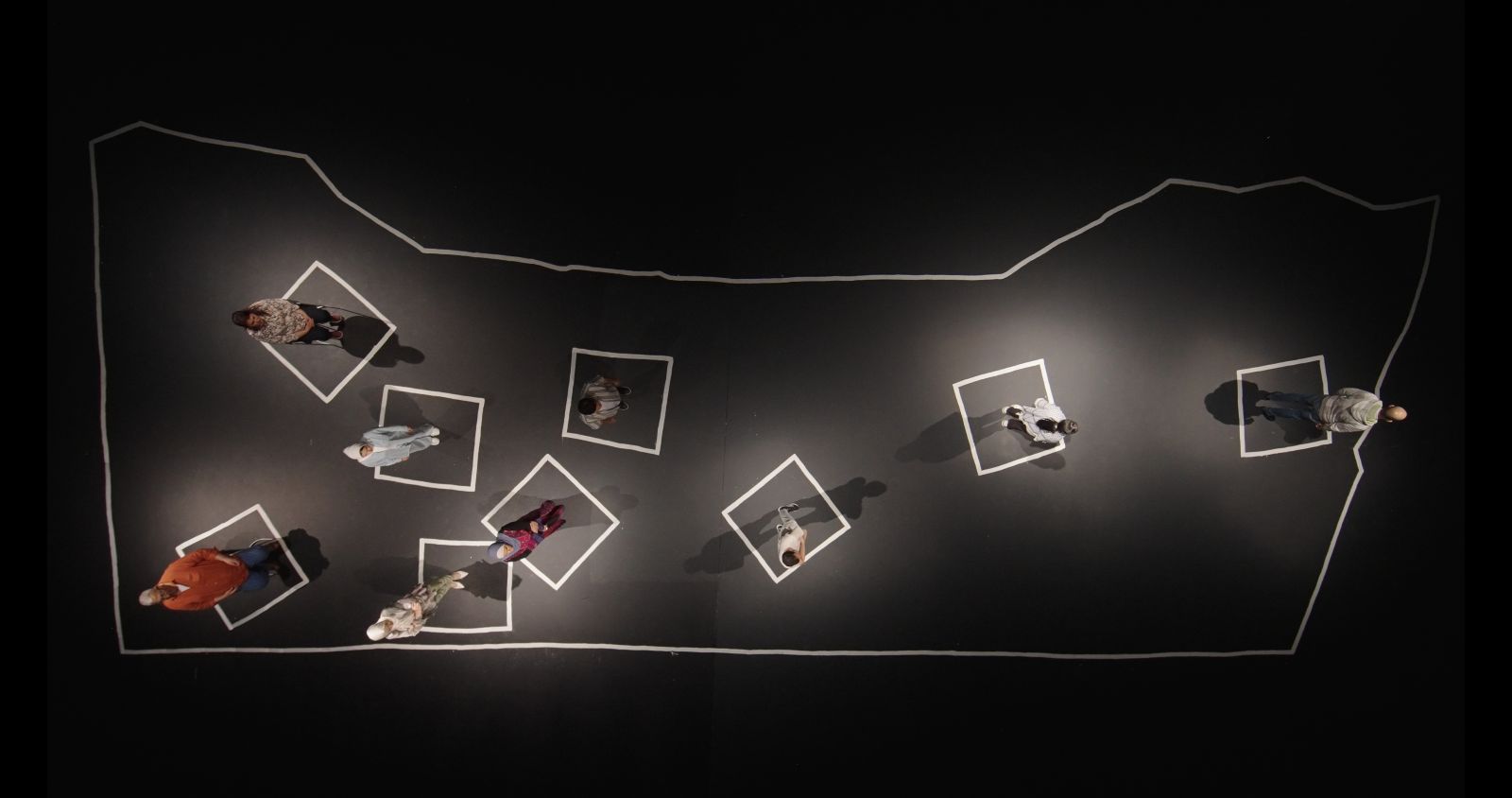 An overhead view of a soundstage covered in a black canvas as nine Palestinians stand in white square outlines that represent their former neighbourhoods and homes.