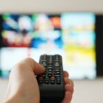 A hand holds a remote control in the air and points it towards a TV in the background. The TV is out of focus.