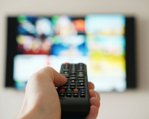 A hand holds a remote control in the air and points it towards a TV in the background. The TV is out of focus.