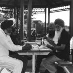 A black and white photo depicts a group of South Asian men wearing turbans play cards on a picnic table at a park in Brampton. One of them looks into the camera with a hint of a smile.