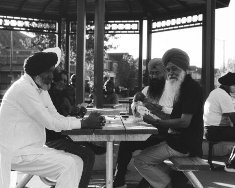 A black and white photo depicts a group of South Asian men wearing turbans play cards on a picnic table at a park in Brampton. One of them looks into the camera with a hint of a smile.