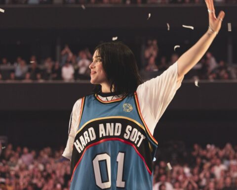 Singer Billie Eilish stands on stage with her hand raised, waiving to fans. She is a 24-year-old white woman with brown hair. She is smiling and wearing a blue jersey with white sleeves that has a black arc that says 'Hard and Soft' in while letters, and the numbers 01 beneath in white.