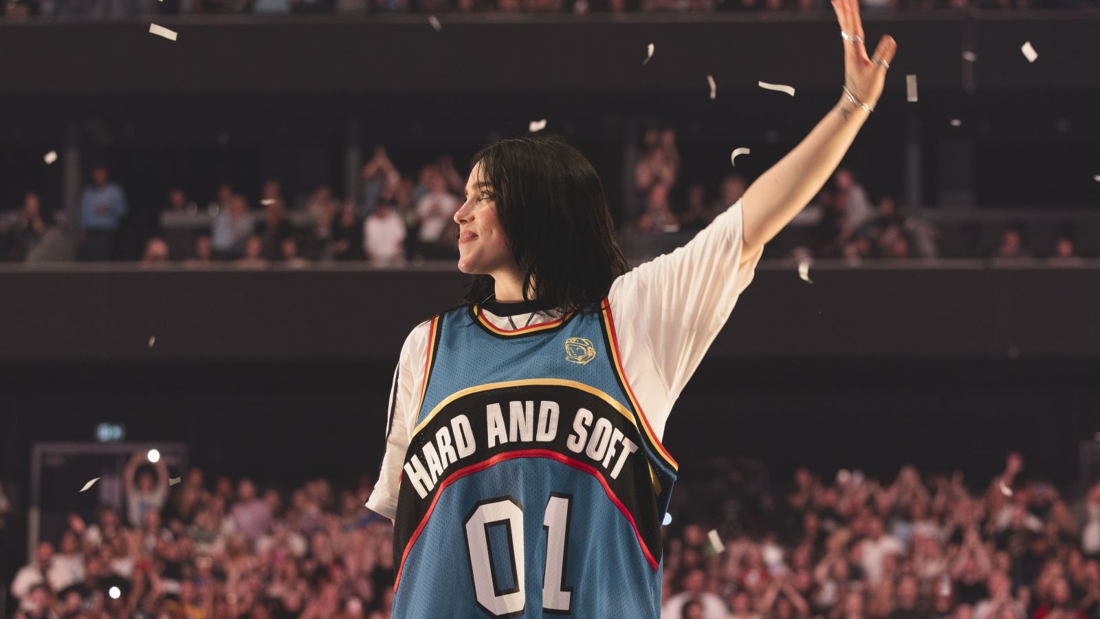 Singer Billie Eilish stands on stage with her hand raised, waiving to fans. She is a 24-year-old white woman with brown hair. She is smiling and wearing a blue jersey with white sleeves that has a black arc that says 'Hard and Soft' in while letters, and the numbers 01 beneath in white.